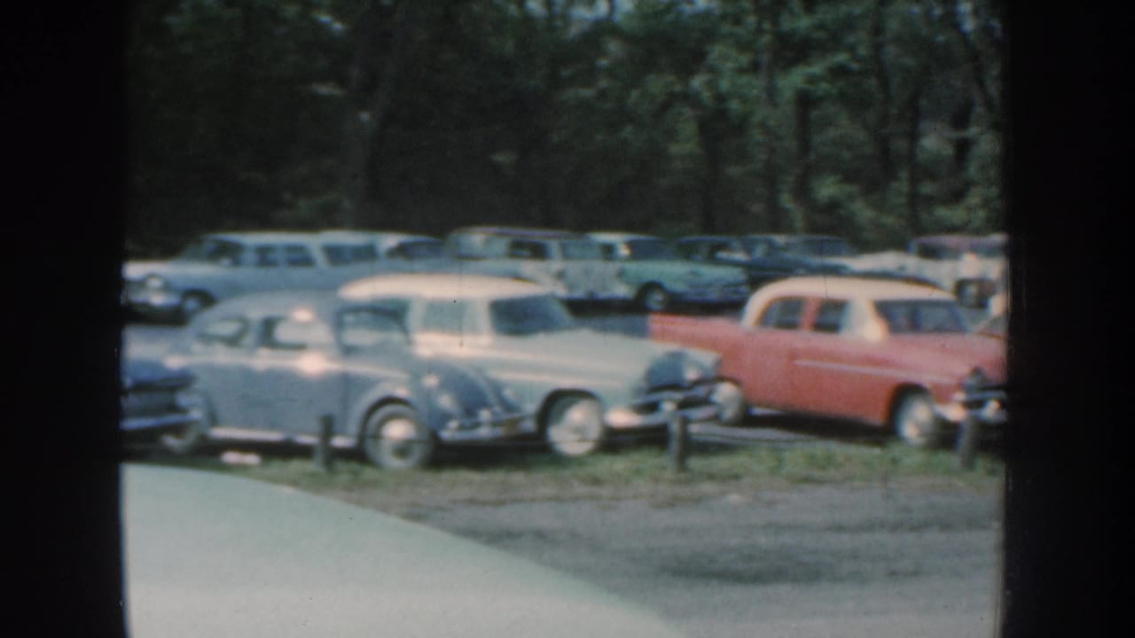 Light-colored vintage cars and a red station wagon parked in a tree-lined lot.