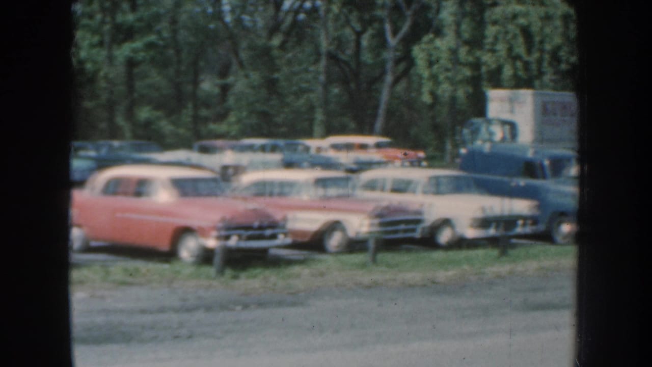 1959 Nebraska frame showing a red station wagon, pale station wagons, and a delivery truck in a busy wooded parking area.