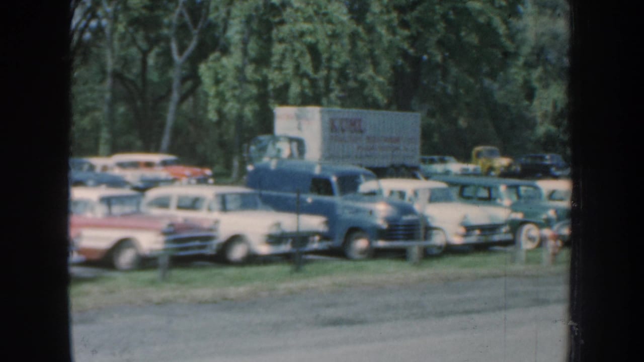 1959 Nebraska 8mm frame with a lettered delivery truck, classic parked cars, and a wooded background in a grassy parking area.
