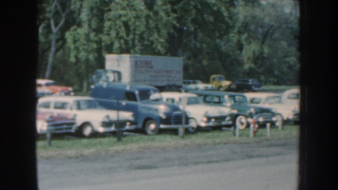1959 Nebraska 8mm frame showing classic cars in a grassy roadside parking lot, with a white box truck and trees in the background.