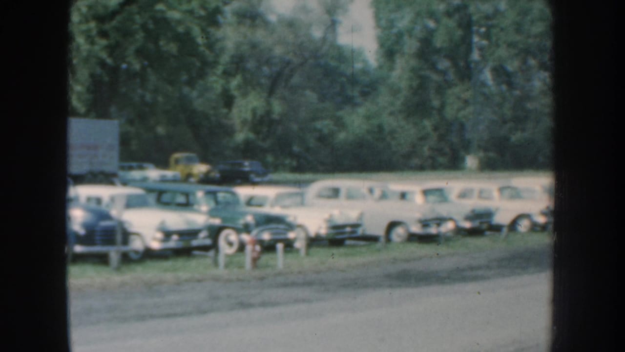 Parked cars and trucks along a grassy roadside lot, seen through soft 8mm film grain.