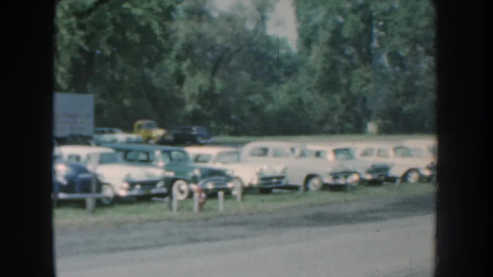 1959 Nebraska roadside view with rows of parked classic cars, a box truck, trees, and open road in soft 8mm color.