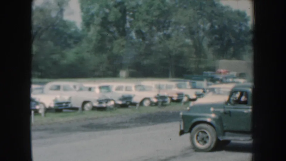 Green truck driving past a 1959 Nebraska roadside parking area with classic cars and trees behind it.