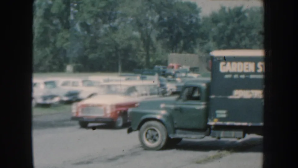 1959 Nebraska frame with a delivery truck bearing garden and poultry lettering passing a lot of parked classic cars.