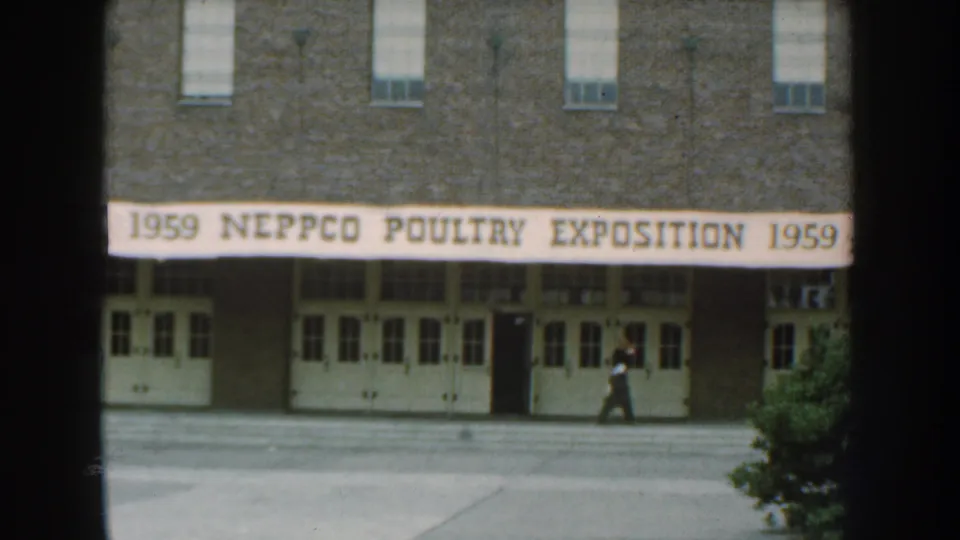 1959 Nebraska home-movie frame showing the NEPPCO Poultry Exposition banner across a brick building as a person walks by.