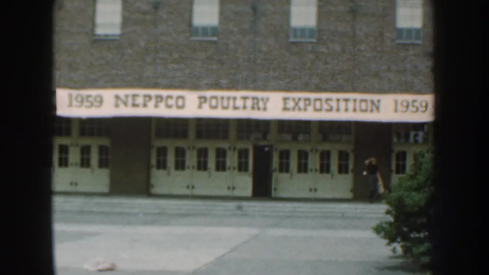 1959 Nebraska home-movie frame of the poultry exposition venue with banner, brick facade, white doors, and a person at the far right.