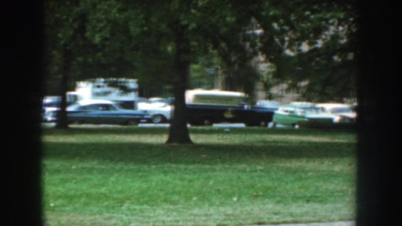 1959 Nebraska home-movie frame showing a grassy lawn, trees, and several parked cars viewed through dark window mullions.
