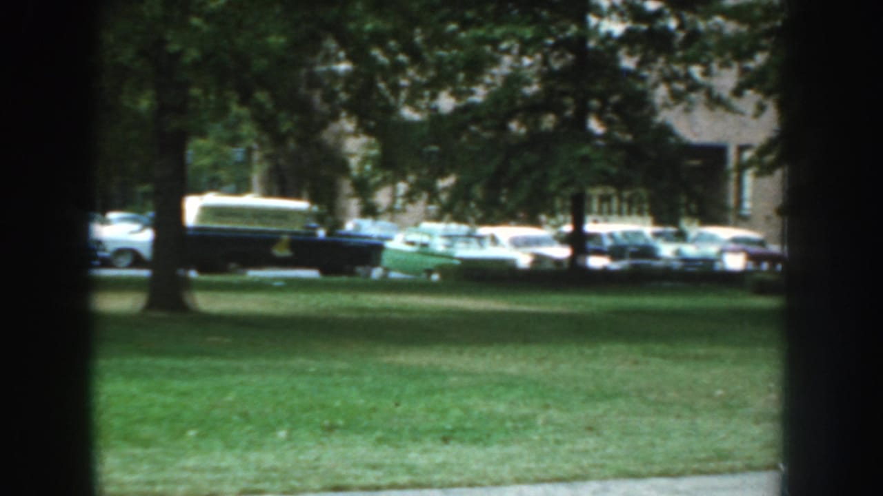 Trees and lawn in foreground with classic cars parked along the road near a brick building.