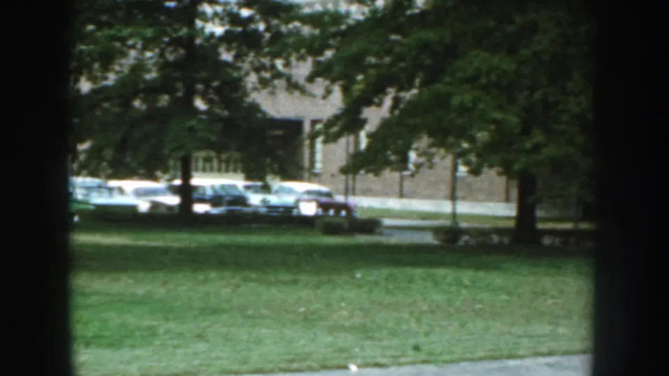 1959 Nebraska frame with parked mid-century cars, a brick building, trees, and lawn seen from inside or behind a window.