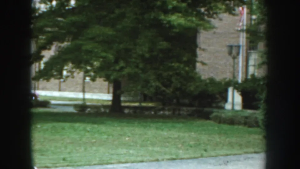 1959 Nebraska home-movie frame showing a lawn with trees, benches, a brick building, and a flagpole at the edge of the scene.