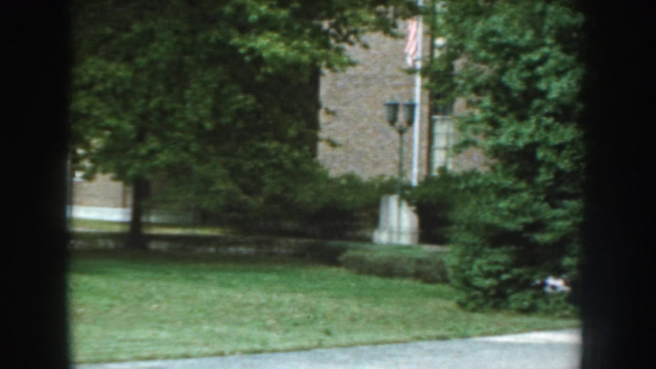 1959 Nebraska 8mm frame showing a brick institutional building beyond lawn and trees, partially obscured by dark vertical framing.