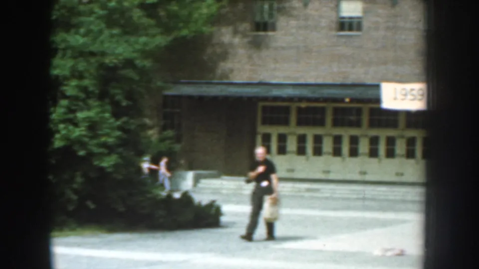 1959 Nebraska 8mm frame with a man carrying a bag across a paved courtyard near trees, brick walls, and an exposition banner edge.