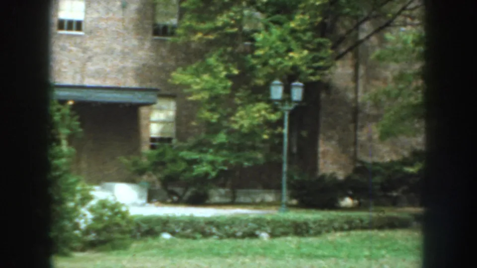 1959 Nebraska exterior frame showing the exposition building grounds with a brick facade, trees, walkway, lawn, and lamppost.