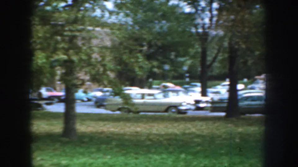 1959 Nebraska home-movie frame showing parked mid-century cars seen through dark tree trunks and green lawn.