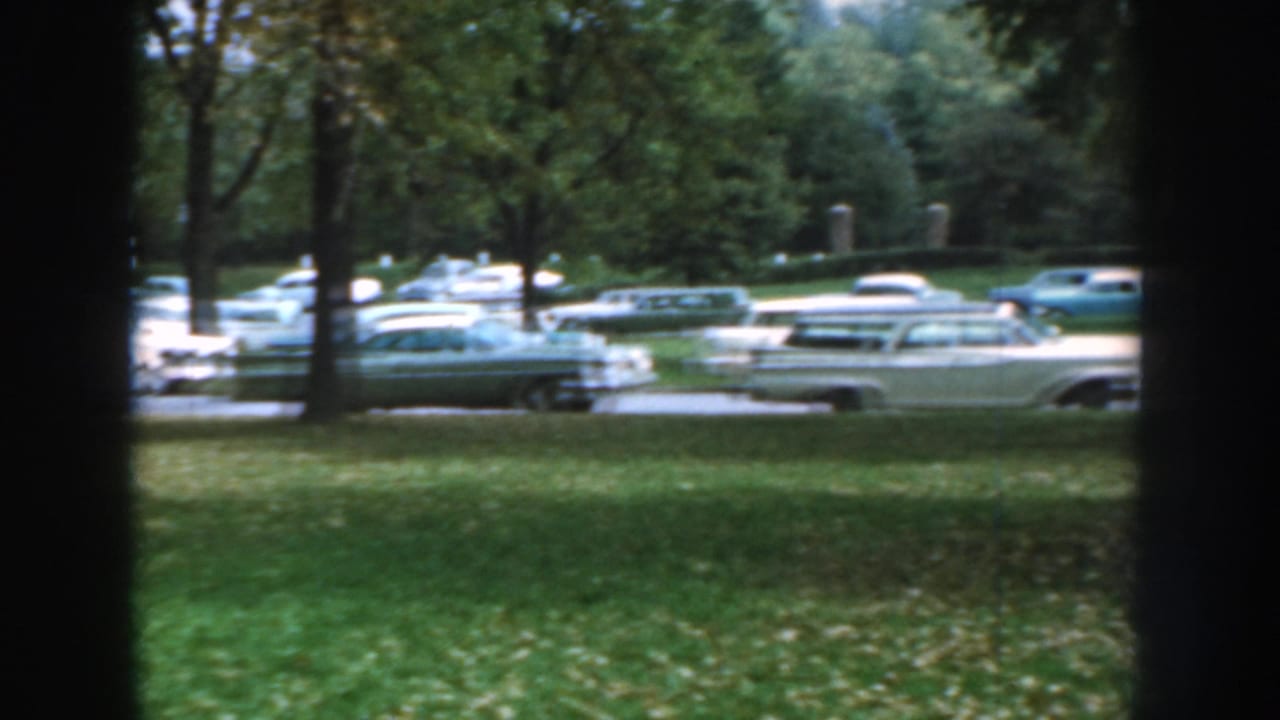 A 1959 Nebraska frame shows cream, blue, and green cars parked in rows beyond a grassy foreground.