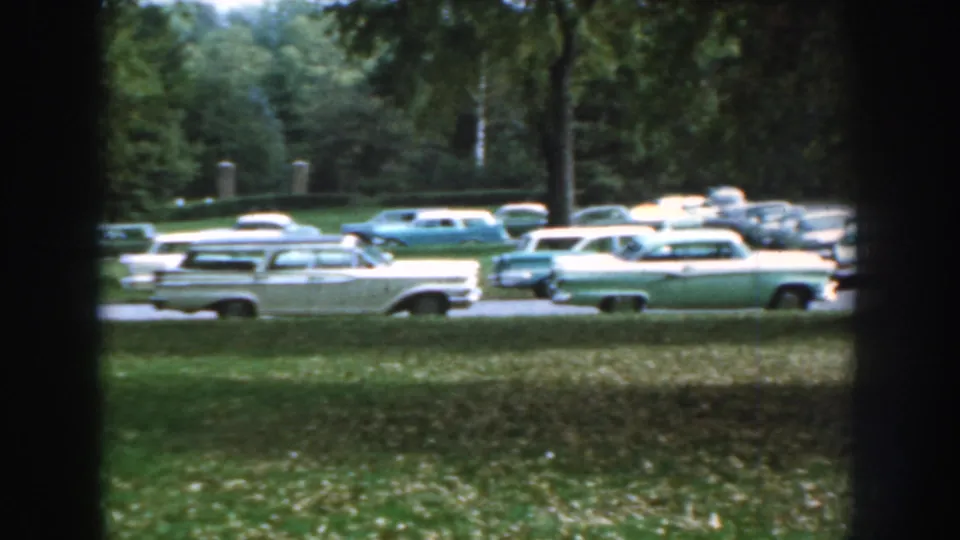 1959 Nebraska home-movie frame with multiple classic cars parked along a shaded roadway or lot edge.