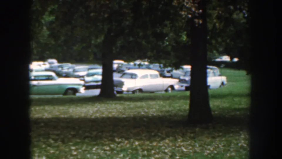 1959 Nebraska home-movie frame showing parked cars beyond trees and grass near the poultry expo grounds.
