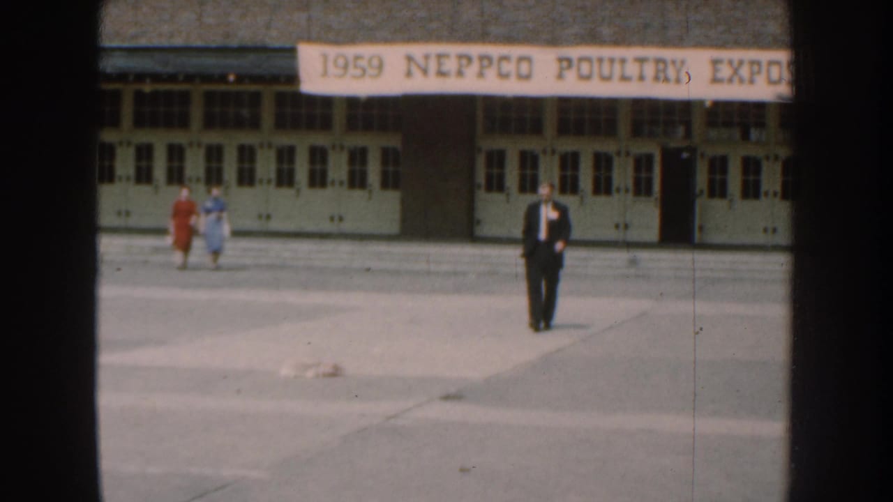 A suited man crosses a paved courtyard in front of a building with a 1959 NEPPCO Poultry Expo banner in Nebraska.