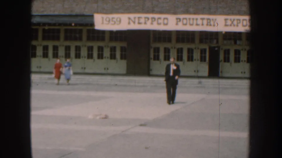 A suited man crosses a paved courtyard in front of a building with a 1959 NEPPCO Poultry Expo banner in Nebraska.