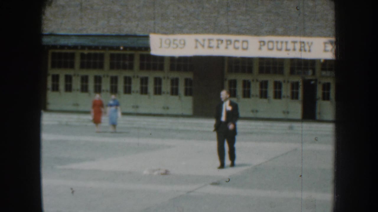 In 1959 Nebraska footage, a suited man walks toward the camera while two women pass behind him under a NEPPCO poultry expo banner.