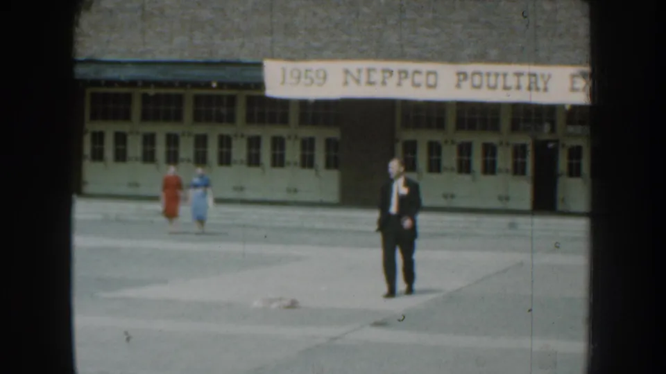 In 1959 Nebraska footage, a suited man walks toward the camera while two women pass behind him under a NEPPCO poultry expo banner.