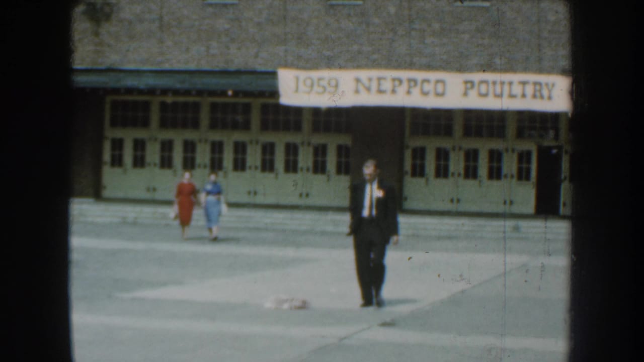 The suited man advances across the paved foreground as two women in red and blue dresses walk near the building behind him.