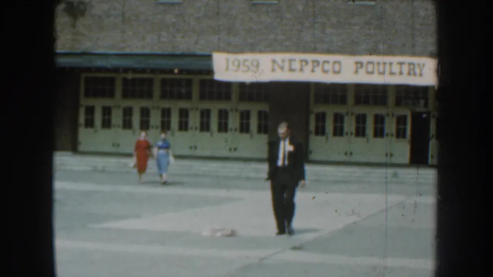 The suited man advances across the paved foreground as two women in red and blue dresses walk near the building behind him.
