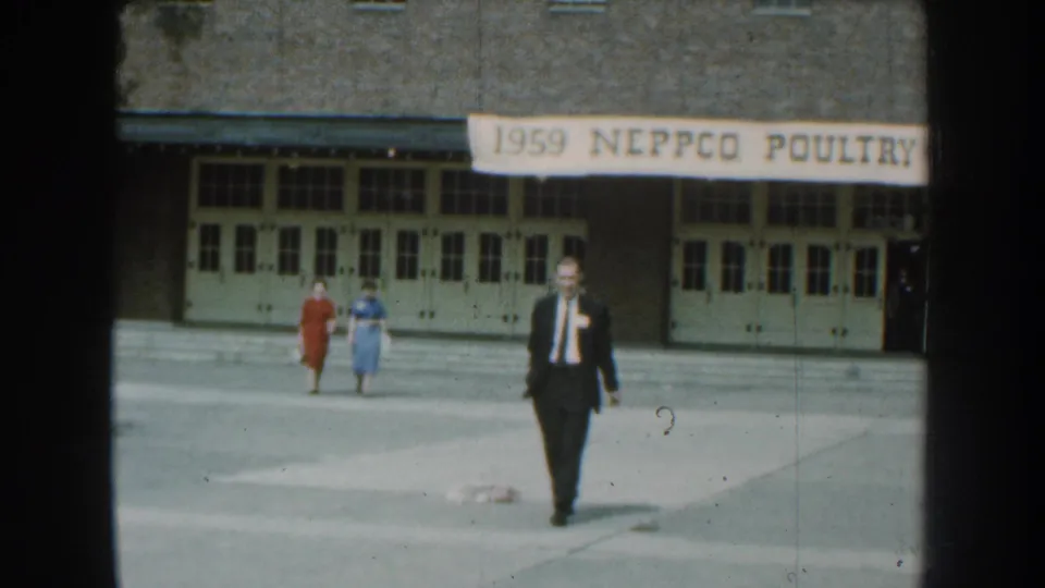 The 8mm frame shows the man closer in the foreground, with the NEPPCO banner and two women still visible behind him.