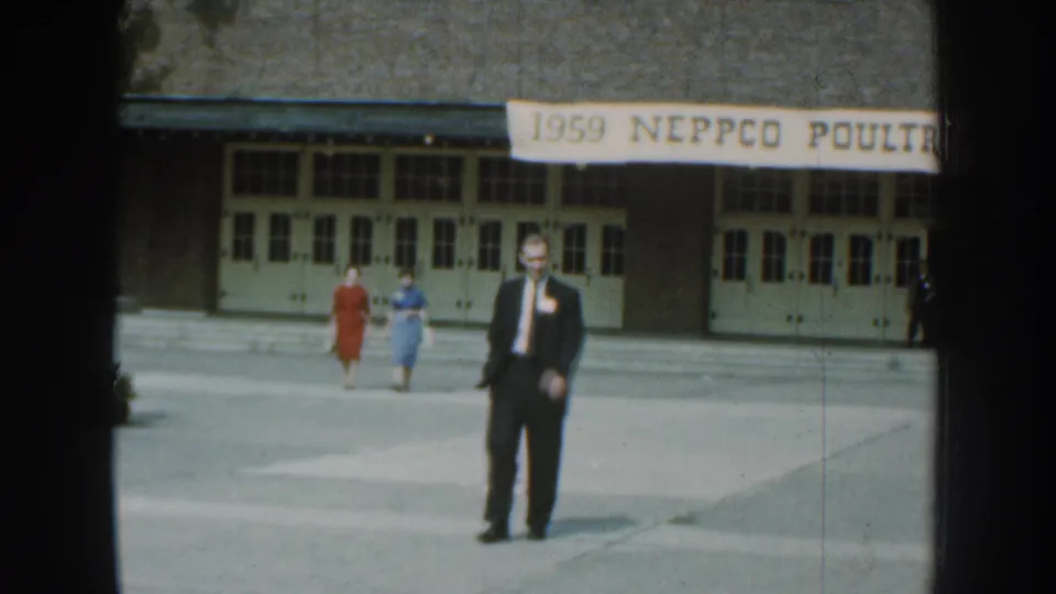 Near the end of the clip, the man in a dark suit stands or slows near the camera before the 1959 NEPPCO poultry expo building.