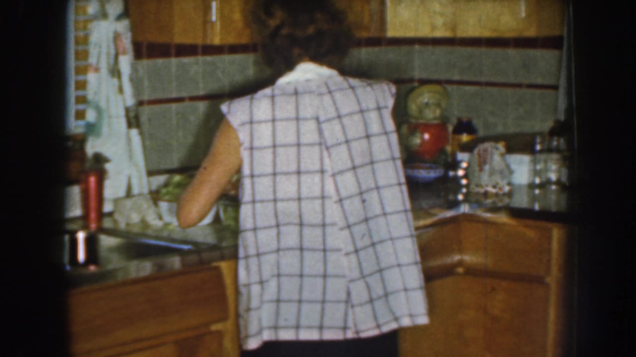In a 1959 Nebraska home kitchen, a woman in a sleeveless plaid top works at a counter near a sink and tiled backsplash.