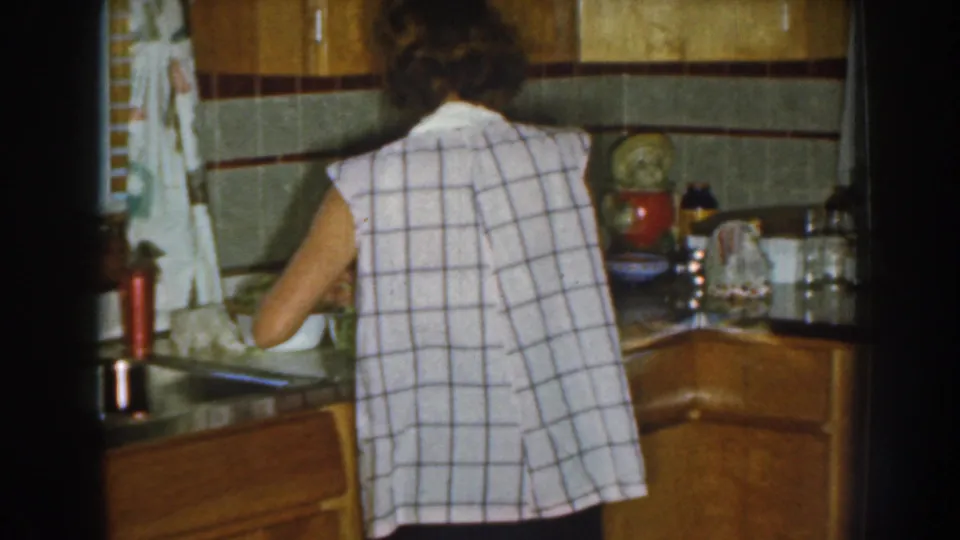 The woman remains turned away while preparing food in a Nebraska kitchen with wood cabinetry, tile and countertop items.