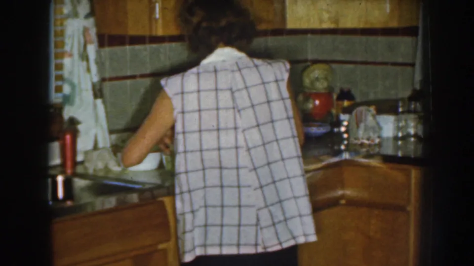A closer 1959 home-movie view shows the woman leaning over the counter or sink area in the same retro Nebraska kitchen.