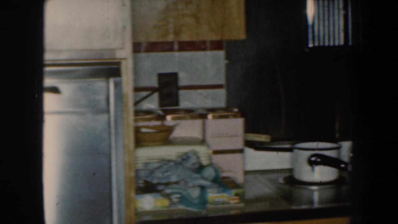 1959 Nebraska home-movie frame showing a stainless refrigerator and a busy kitchen counter with boxes, dishes, and towels.