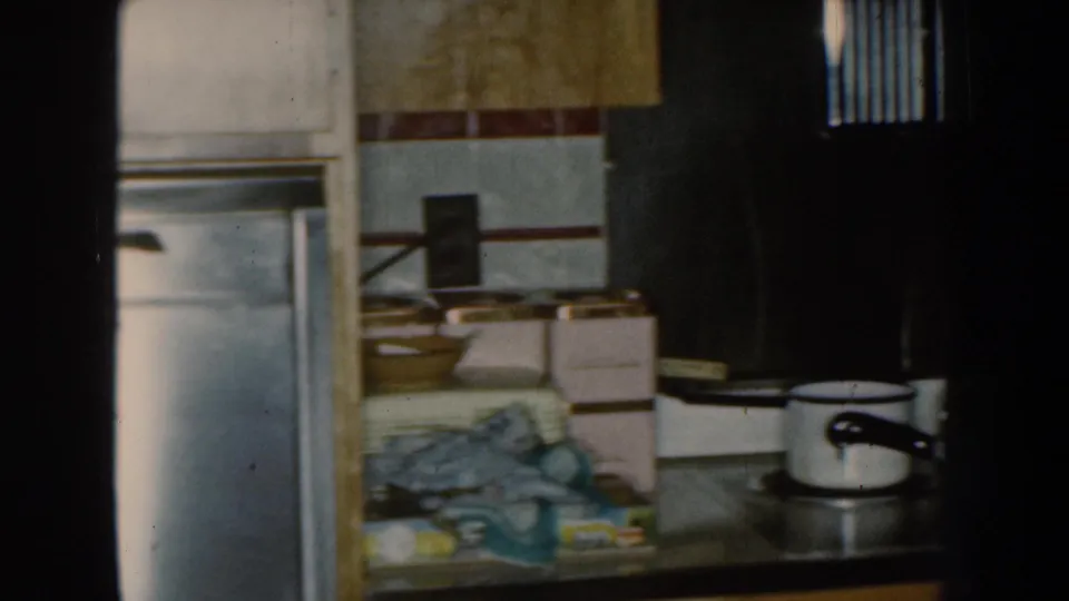 1959 Nebraska home-movie frame showing a stainless refrigerator and a busy kitchen counter with boxes, dishes, and towels.