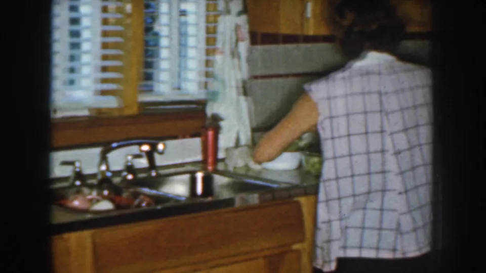 1959 Nebraska kitchen frame with a woman seen from behind working at the sink beside wood cabinets and white blinds.