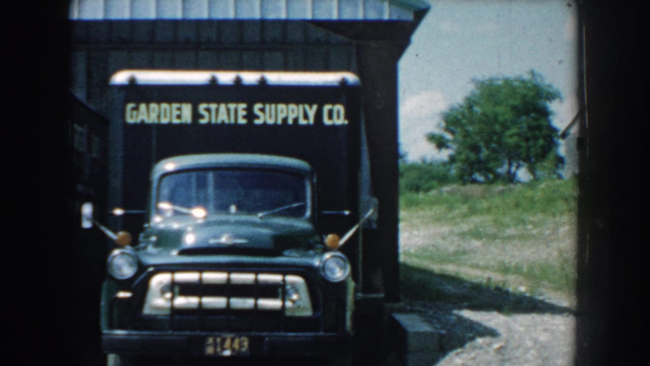 1959 Nebraska 8mm frame showing a dark Garden State Supply Co delivery truck facing the camera near a corrugated service structure.