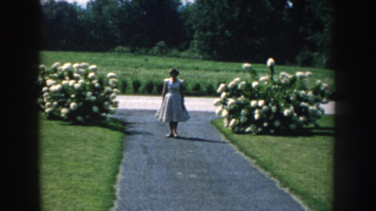 1959 Nebraska 8mm frame showing a female figure on a paved garden path, flanked by white hydrangeas and green lawns.