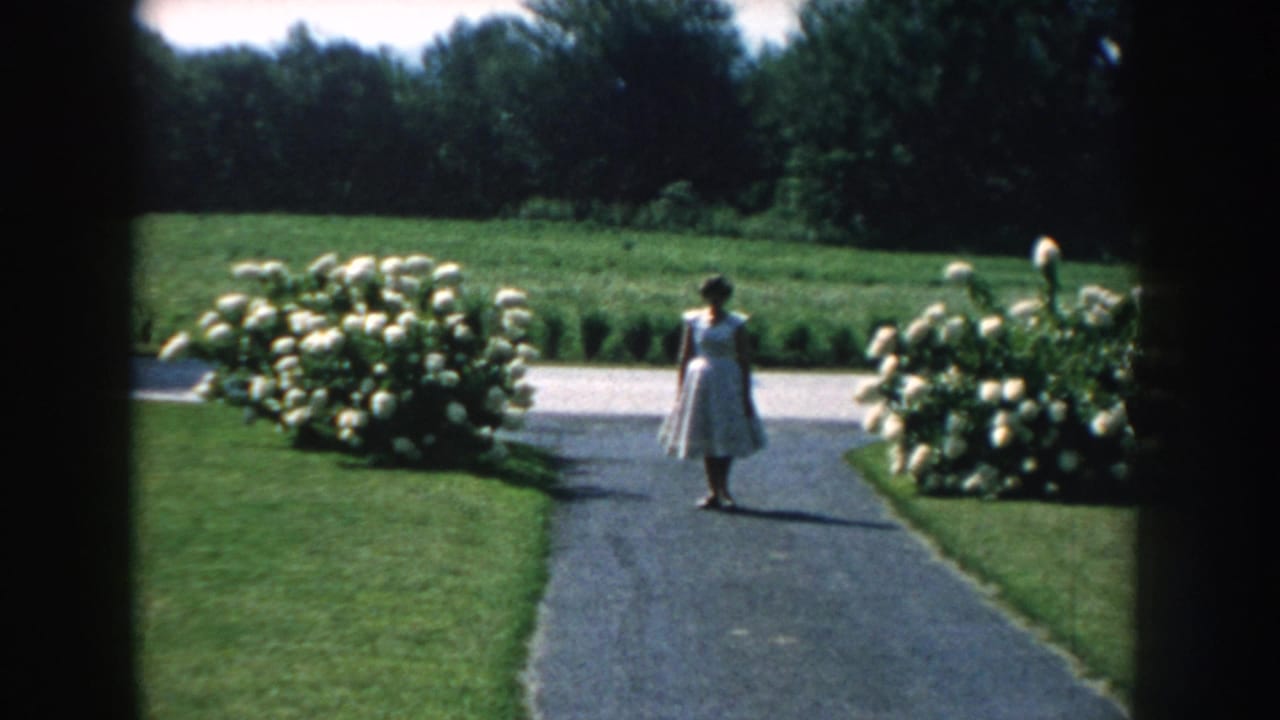 The 1959 Nebraska garden view holds on the figure in a pale dress, framed by lawns, hydrangeas, and distant trees.