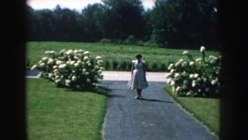 The 1959 Nebraska garden view holds on the figure in a pale dress, framed by lawns, hydrangeas, and distant trees.