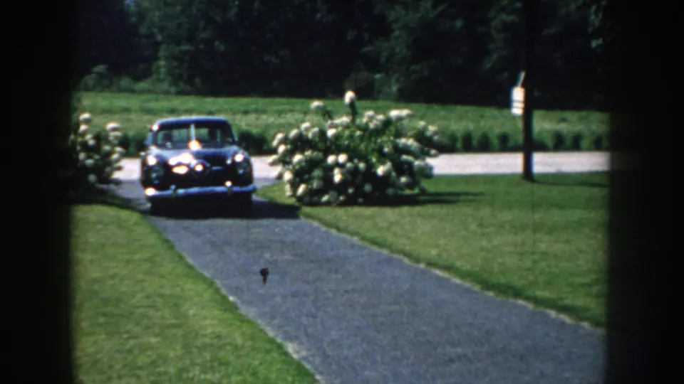 A dark late-1950s-style car appears on the path in the Nebraska garden scene, with hydrangeas and trimmed grass visible.