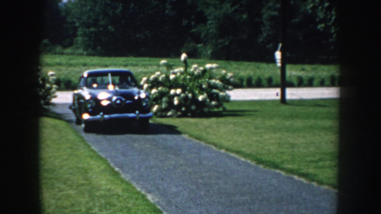1959 Nebraska home-movie frame showing a dark mid-century sedan entering a paved driveway through shaded foreground edges.