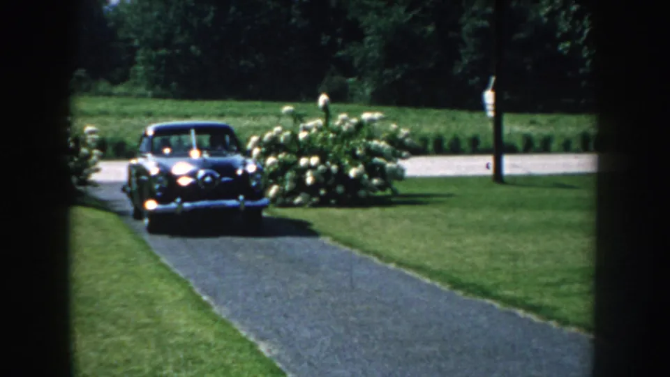 1959 Nebraska home-movie frame showing a dark mid-century sedan entering a paved driveway through shaded foreground edges.