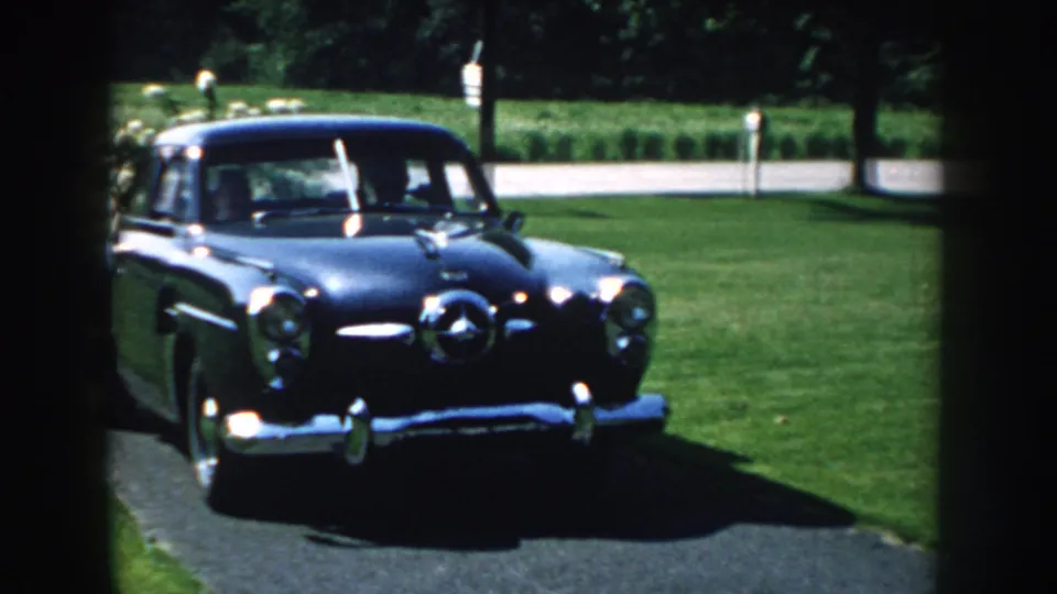 Close 1959 home-movie view of a dark sedan with chrome details nearing the camera on a paved driveway.
