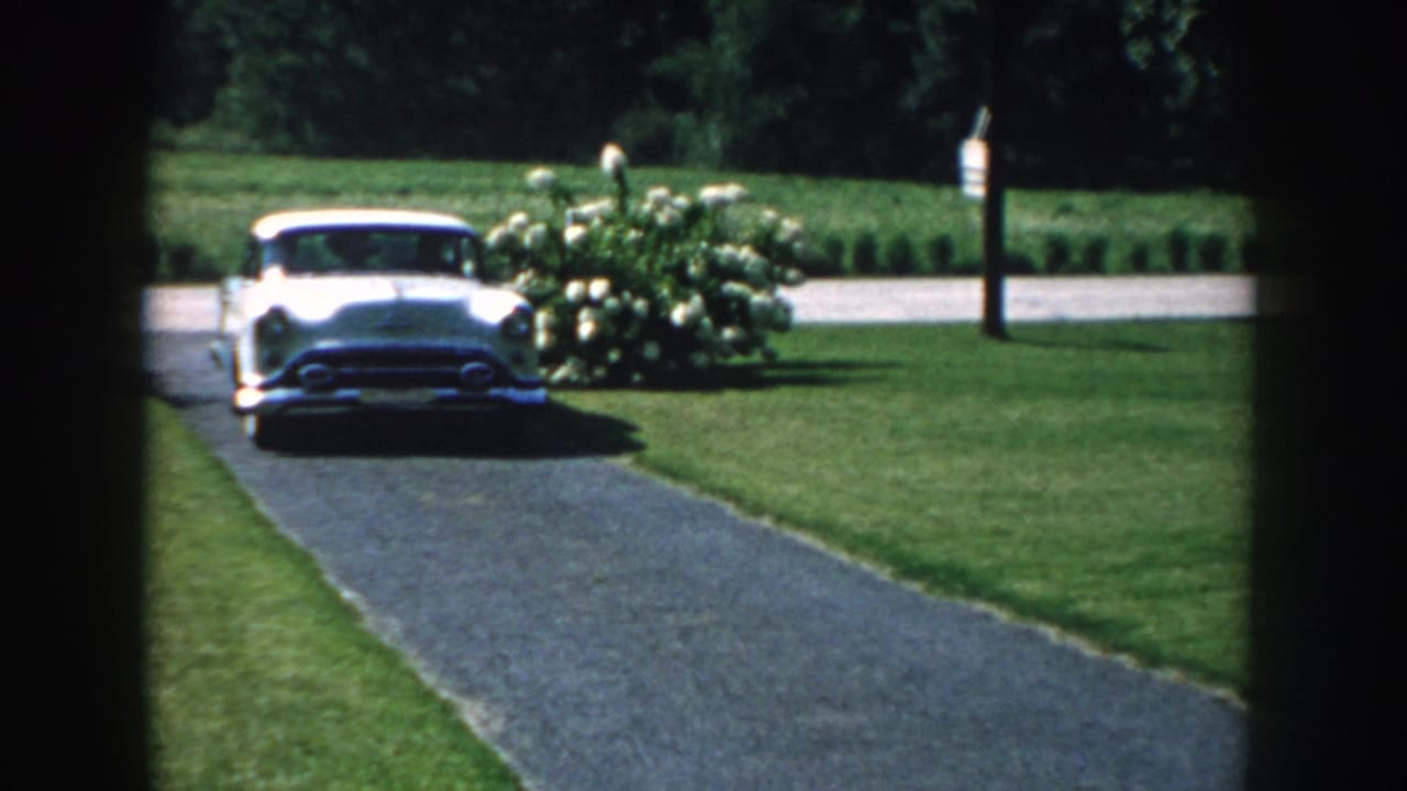 A white 1950s sedan drives up a paved driveway in a Nebraska home-movie scene, framed by dark foreground edges and green lawn.