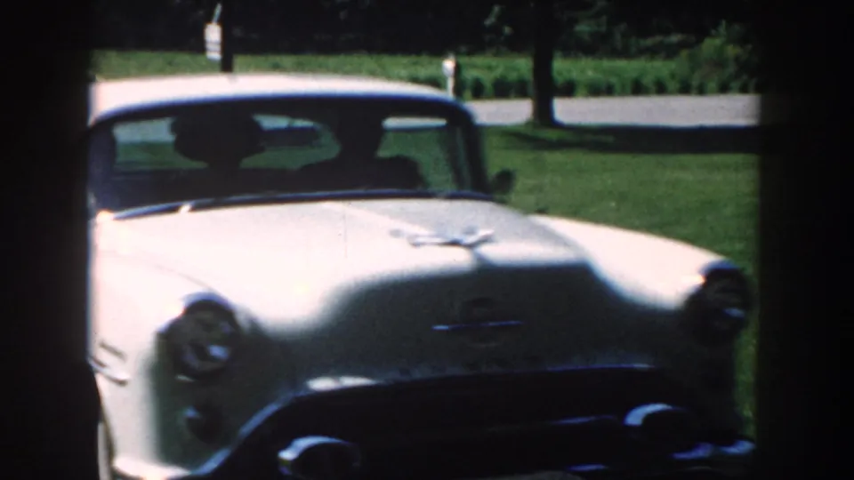 A close, soft-focused front view shows the white 1950s sedan filling much of the 8mm frame as it reaches the driveway near the camera.