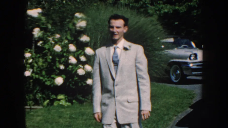 An adult man in a light suit and tie stands on the lawn near flowering shrubs, with the white 1950s sedan parked behind him in Nebraska footage.