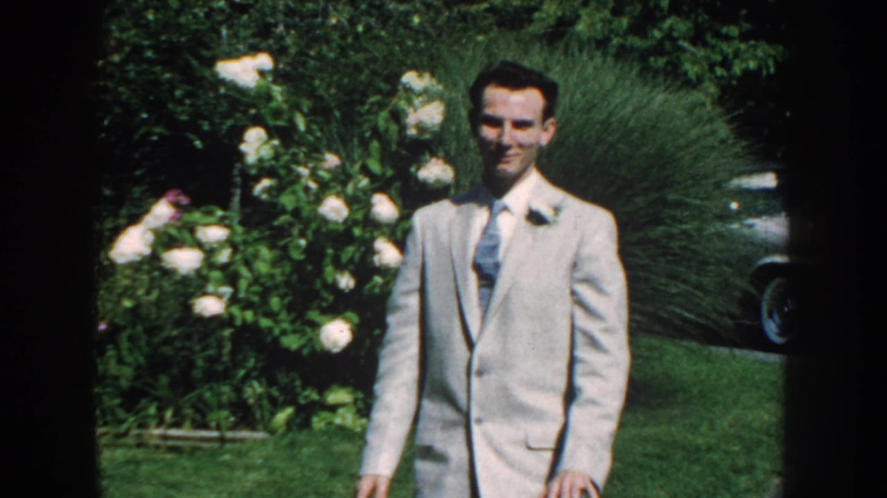 In Nebraska 1959 home-movie footage, a man in a light suit with boutonniere walks through a green backyard garden.