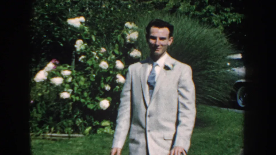 In Nebraska 1959 home-movie footage, a man in a light suit with boutonniere walks through a green backyard garden.