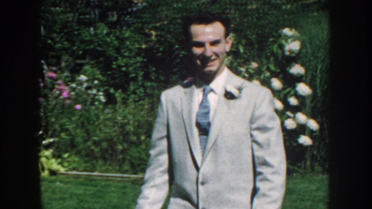 Smiling man in a light suit stands or walks in front of garden hedges and flowers.