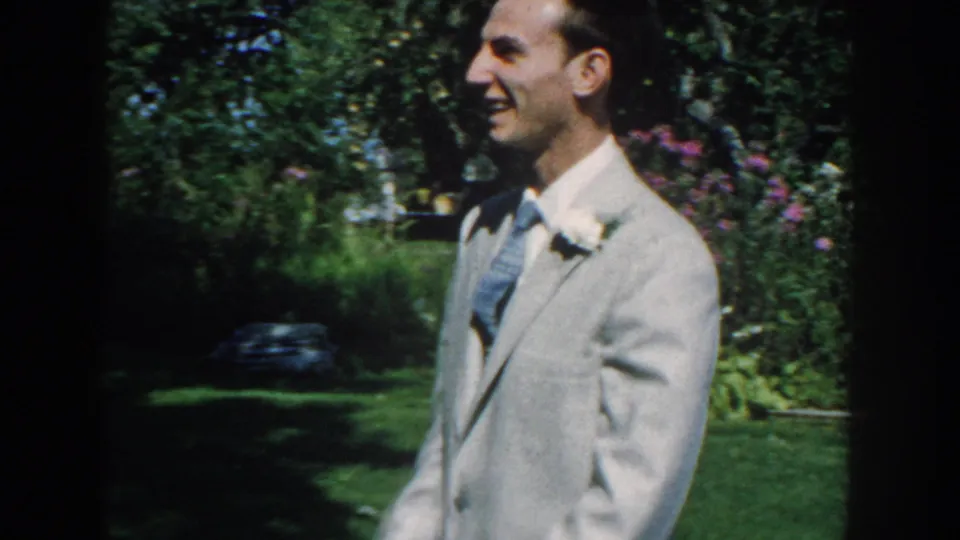 A side view shows the suited man smiling in a leafy Nebraska backyard garden, with flowers and lawn visible around him.